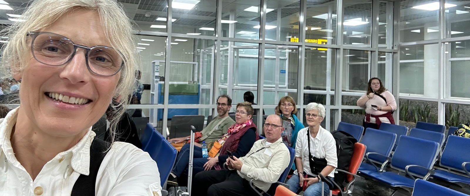 Das Foto zeigt Benedicte. Im Selfie-Modus hat sie das Bild aufgenommen. Sie lacht in die Kamera. Im Hintergrund erkennt man weitere Personen der Reisegruppe. Sie sitzen in einer Abflughalle des Flughafens in Kigali.